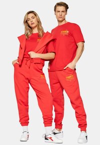 Tuta in cotone rosso composta da felpa con cappuccio e pantaloni jogger, entrambi con stampe grafiche gialle. I pantaloni jogger hanno polsini elastici e tasche.