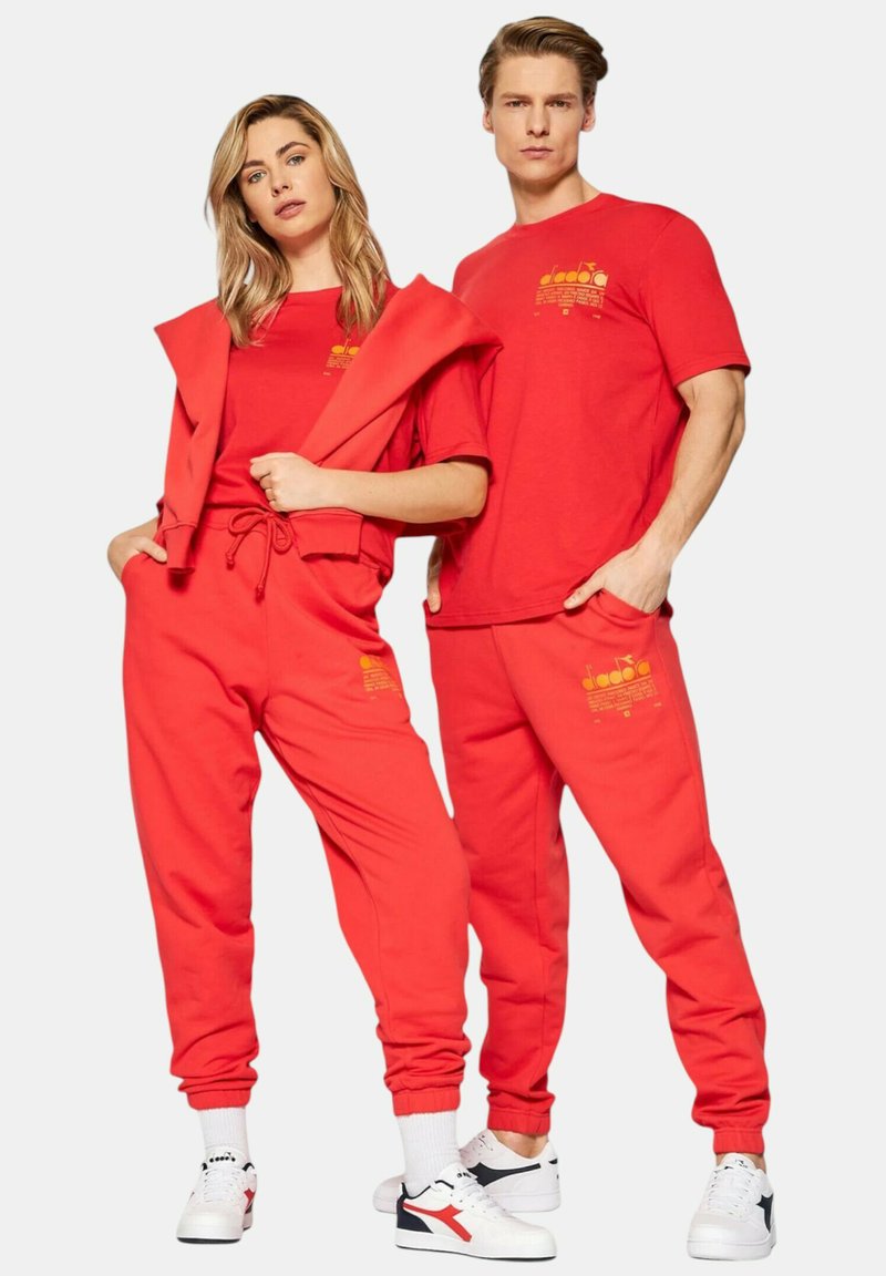 Tuta in cotone rosso composta da felpa con cappuccio e pantaloni jogger, entrambi con stampe grafiche gialle. I pantaloni jogger hanno polsini elastici e tasche.