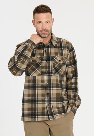 Middle-aged man with short hair wearing a brown and beige plaid button-up shirt and tan pants, touching his collar with one hand.