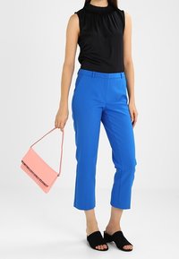 Top nero senza maniche con scollatura arricciata, pantaloni blu brillante a vita alta, borsa rosa con design a contrasto e scarpe open-toe nere senza lacci.
