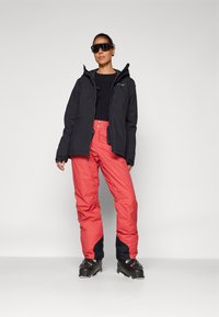 Veste noire isolée avec capuche, assortie à un pantalon de ski rouge vif avec des accents noirs. Le modèle porte de lunettes de soleil élégantes et des chaussures de ski.