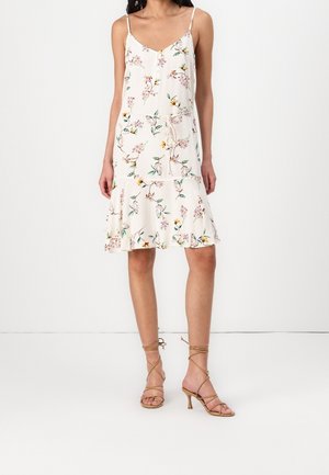 Vrouw draagt een mouwloze witte jurk tot op de knie met bloemenprint en beige, hooggehakte sandalen met bandjes, staand tegen een witte achtergrond.