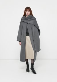 Grijze wollen jas met een oversized kraag, gelaagd ontwerp en lange mouwen. Gecombineerd met een lichtbeige rok en zwarte hakken.