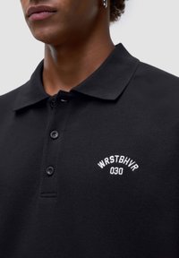 Schwarzes Poloshirt aus strukturiertem Stoff, mit einer Knopfleiste mit drei Druckknöpfen und einem weißen gestickten Logo auf der linken Seite.