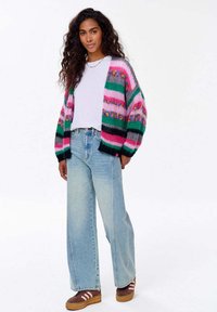 Barevný pletený cardigan s růžovými, zelenými a černými pruhy, nošený přes bílou košili. Kombinován s modrými džíny s širokými nohavicemi a hnědými botami na platformě.