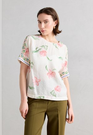 Donna che indossa una camicetta bianca con stampa di fiori rosa e maniche con motivo verde, abbinata a pantaloni verde oliva, in piedi davanti a uno sfondo neutro.