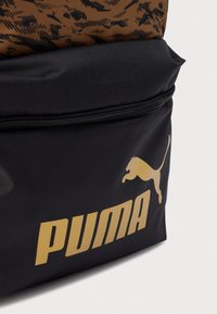 Μαύρη βάση σακιδίου με καφέ διακοσμημένη κορυφή, με χρυσό λογότυπο "PUMA" και γραφικό ενός πηδώντας πούμα. Ορατό φερμουάρ κλεισίματος.