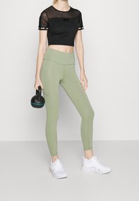 Černý síťovaný crop top s logem, v kombinaci s olivově zelenými legínami s vysokým pasem a bílými sportovními botami. Drží černou kettlebell.