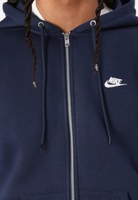 Marineblå hettejakke med dobbel snor, sølvglidelås og liten hvit Nike-logo på venstre bryst. Mykt stoff med en jevn tekstur.