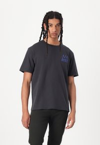 Short-sleeve black t-shirt featuring a blue crown logo on the left chest. The fabric appears soft with a relaxed fit. Worn with dark pants.