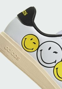 Bijele tenisice s crnim detaljima; sadrže tri grafike s osmijehom u žutoj i crnoj boji; teksturirana bež guma potplat s utisnutim logotipom.