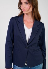 Blazer blu navy in tessuto liscio con chiusura a un bottone, revers a punte e due tasche frontali, abbinato a una maglietta bianca a V.