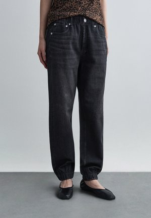 Personne portant un jean noir avec bords élastiques, des ballerines noires et un haut marron à imprimé léopard, debout sur un sol gris devant un fond uni.