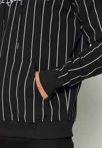 Musta huppari, jossa on pystysuorat valkoiset raidat, edessä tasku, ribbaus hihansuissa ja helmoissa. Nyöritön huppu, jossa on metalliset päället.