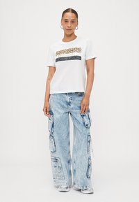 T-shirt blanc à manches courtes avec motif animal et texte doré ; associé à un jean cargo large bleu clair avec poches et finition délavée.