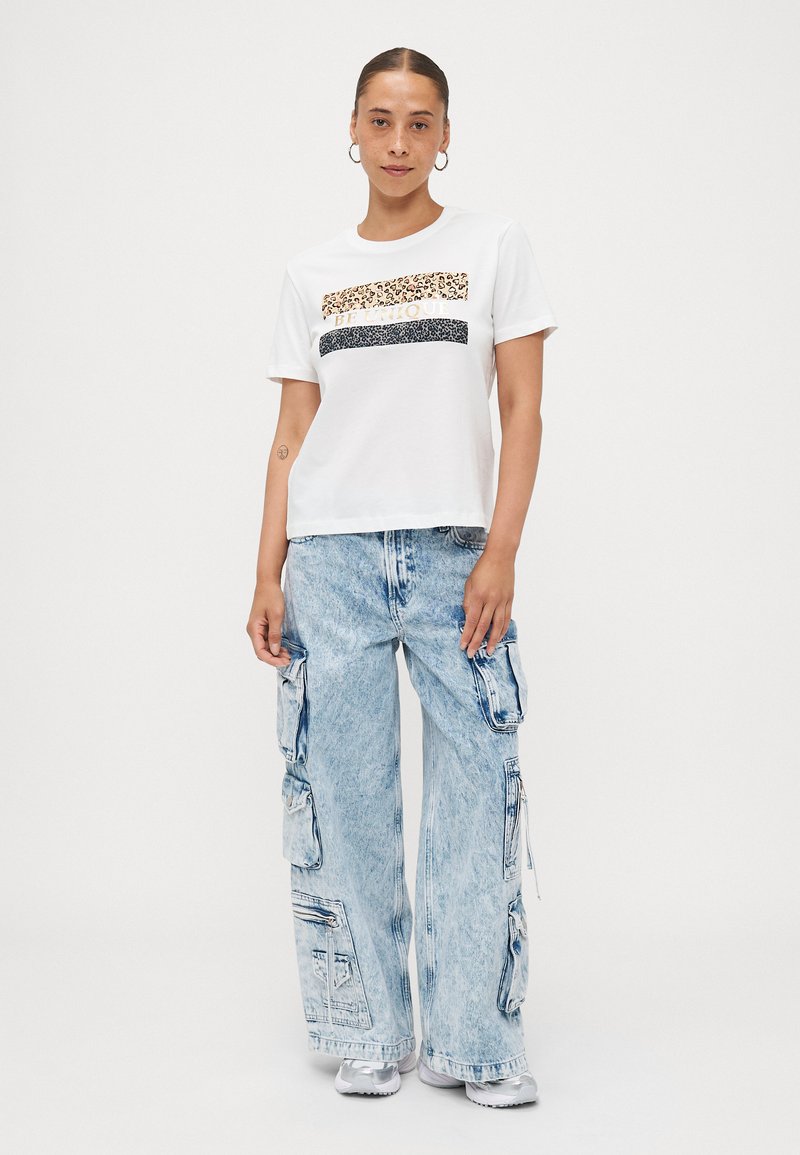 Weißes T-Shirt mit kurzen Ärmeln, Tierdruck und goldener Schrift; kombiniert mit hellblauen weiten Cargo-Jeans, die Taschen und einen blassen Look aufweisen.