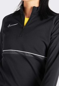Sudadera deportiva negra de media cremallera con un forro interior amarillo, presenta una franja blanca y una banda texturada negra en el pecho. El logo de Nike es visible.