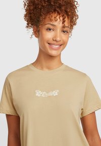 Beige short-sleeve t-shirt made of soft fabric featuring a floral logo design on the chest in white. Smooth texture, casual fit.
