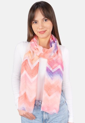 Young woman with long brown hair wearing a white top and light blue jeans, smiling, with a pink and purple zigzag patterned scarf.