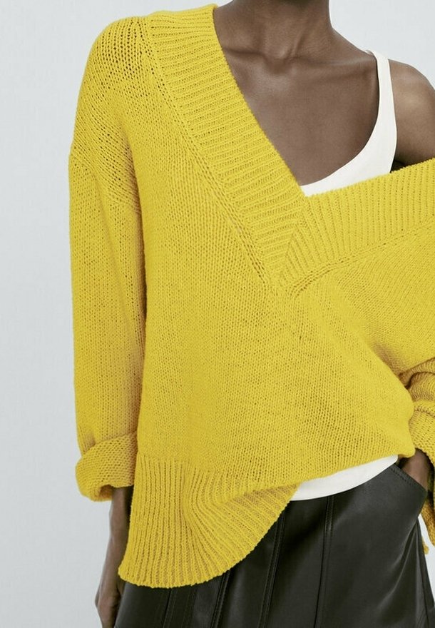 Personne portant un pull en maille jaune ample par-dessus un débardeur blanc et une jupe en cuir noire, avec une main dans la poche.