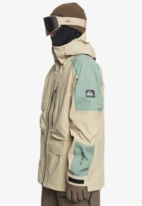 Giacca impermeabile beige e verde con molte tasche, polsini regolabili e frontale con zip. Presenta un logo sulla manica.
