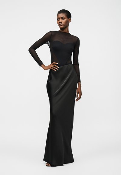 Γυναίκα που στέκεται φορώντας μαύρο διαφανές τοπ με μακριά μανίκια και μαύρη σατέν μακριά φούστα, ποζάροντας με το ένα χέρι στη μέση της μπροστά σε λευκό φόντο.