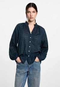 Mörkblå skjorta med knappar och volangkrage, långa ärmar och en subtil mönstrad textur, kombinerad med ljusa denimjeans.