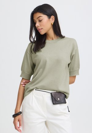 Kobieta z długimi ciemnymi włosami nosi oliwkowozieloną koszulkę z krótkim rękawem, białe spodnie, czarną torebkę na pasku, złote kolczyki i czarną bransoletkę na białym tle.