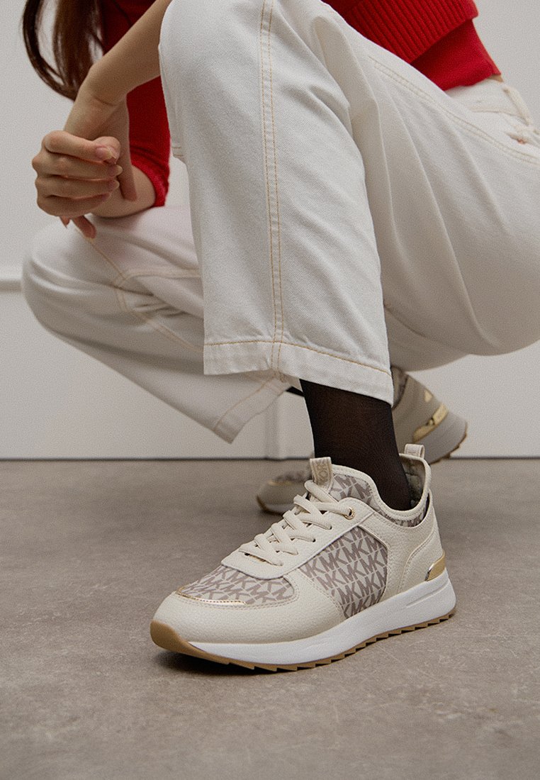 Beige und cremefarbene Sneaker mit strukturiertem Finish, versehen mit einem Monogrammmuster, goldenen Akzenten und einer weißen Sohle, dargestellt auf einem ausgewählten Hintergrund.