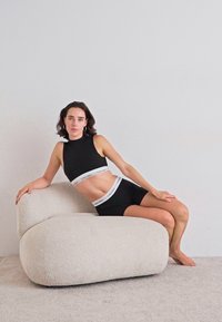 Žena u crnom crop topu i šortsevima brenda Calvin Klein leži na modernoj off-white teksturiranoj fotelji nasuprot jednostavne zida.