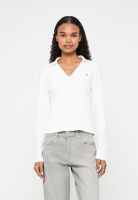 Polo a maniche lunghe bianco con scollo a V e un piccolo logo ricamato sul petto sinistro. Tessuto morbido con vestibilità rilassata. Abbinato a jeans grigi.