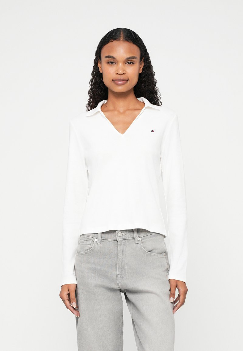 Polo a maniche lunghe bianco con scollo a V e un piccolo logo ricamato sul petto sinistro. Tessuto morbido con vestibilità rilassata. Abbinato a jeans grigi.