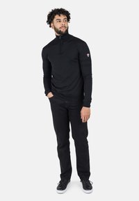 Pullover nero lavorato a maglia con colletto alto e zip a un quarto. Maniche lunghe con polsini aderenti. Indossato con pantaloni neri e scarpe nere.