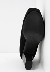 Vue de la semelle d'une chaussure à talon en daim noir avec un bout arrondi, inclinée contre un mur d'angle blanc et le sol.