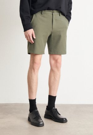 Mężczyzna ubrany w oliwkowo-zielone, dopasowane szorty, czarną koszulę z długim rękawem, czarne skarpetki i czarne skórzane eleganckie buty stoi na jasnej podłodze.