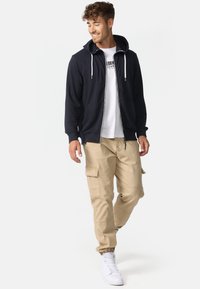 Marineblå hettegenser med glidelås og hvite snorer, lagd over en hvit t-skjorte med grafisk trykk, kombinert med beige cargobukser og hvite joggesko.