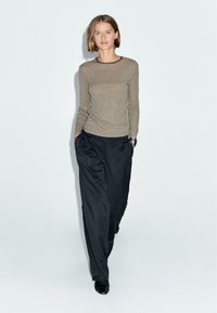 Giovane donna che cammina, indossando una maglietta a maniche lunghe beige e nera a righe, pantaloni neri larghi e scarpe nere su uno sfondo bianco uniforme.
