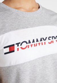 Šedé bavlněné tričko s bílým vodorovným pruhem, na kterém je černý a červený text "TOMMY SPORT", doplněné logem v levém rohu.