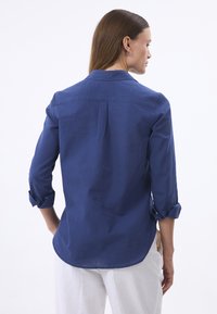 Marineblaues Langarmhemd mit umgeschlagenen Bündchen, glatter Textur und leichtem Glanz. Verfügt über eine Rückenfalte und einen runden Saum.