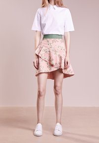 Žena v bílé košili s krátkým rukávem, růžové květinové asymetrické sukni se zeleným pasem a bílých teniskách, stojící na růžové podlaze.