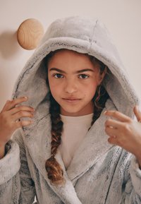Accappatoio in morbido tessuto grigio con ampio cappuccio, caratterizzato da una texture soffice. La persona che lo indossa ha i capelli intrecciati e sta sistemando l'accappatoio. Gancio in legno montato a parete visibile.