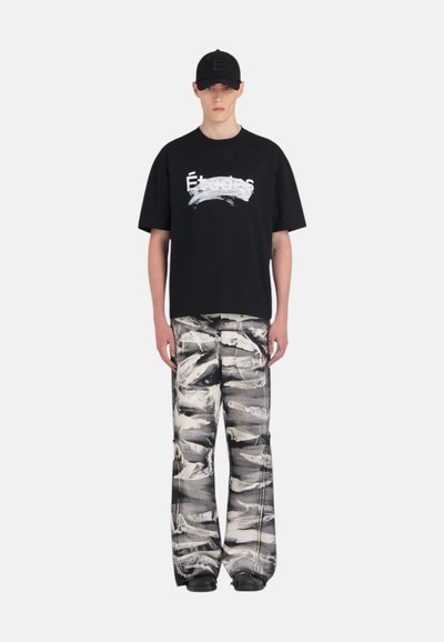 T-shirt oversized noir avec un graphique en traits de pinceau blancs, associé à un pantalon à jambes larges avec un motif abstrait noir et blanc. Casquette noire portée.