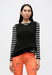 Juodas rankovių megztinis dėvimas ant juodos ir baltos dryžuotos ilgomis rankovėmis marškinio, derinamas su oranžinės spalvos raštuotomis kelnėmis su išpjovomis šonuose.