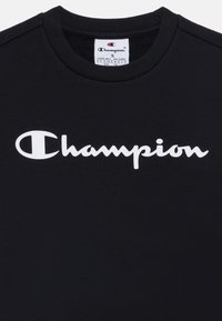 Camiseta de algodón negra con un logo blanco de "Champion" en cursiva. Cuello redondeado y mangas cortas con una textura suave. Etiqueta de tamaño visible.