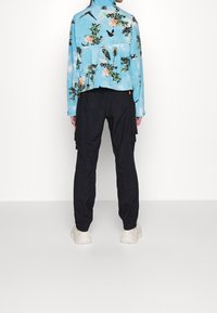 Person som bär en blå jacka med blommor och fågeltryck, svarta cargo-byxor och vita sneakers, står framför en enkel vit bakgrund.