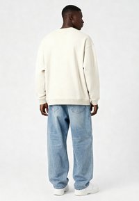 Sudadera de gran tamaño en color beige claro, hecha de una tela suave, combinada con jeans de mezclilla azul claro holgados y zapatillas blancas. Diseño simple y casual.