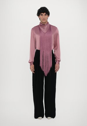 Langärmlige, seidig-mauve Bluse mit Fransenverzierung und hohem Kragen, kombiniert mit hochgeschnittenen schwarzen Hosen und hellen Schuhen.