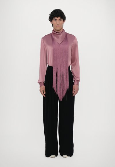 Blusa de seda en tono malva con mangas largas, detalles de flecos y cuello alto, combinada con pantalones negros de talle alto y zapatos de color claro.