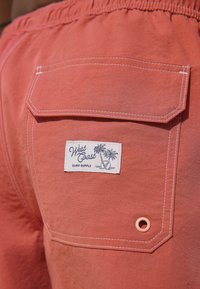 Korall színű úszónadrág rugalmas derékkal, hátul egy zsebbel, amelyen egy fehér címke található, rajta a "West Coast Surf Supply" felirat és egy pálmafás grafika.