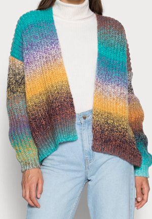 Mujer con cárdigan de punto multicolor con franjas en degradado sobre un jersey blanco de cuello alto y vaqueros azul claro, con las manos relajadas a los lados.